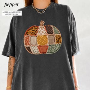 May include: A charcoal grey short-sleeve t-shirt featuring a central graphic of a pumpkin composed of various fabric-style patches in shades of orange, brown, green, and cream. The graphic includes text that reads pepper and NOT REAL EMBROIDERY OR PATCHWORK.