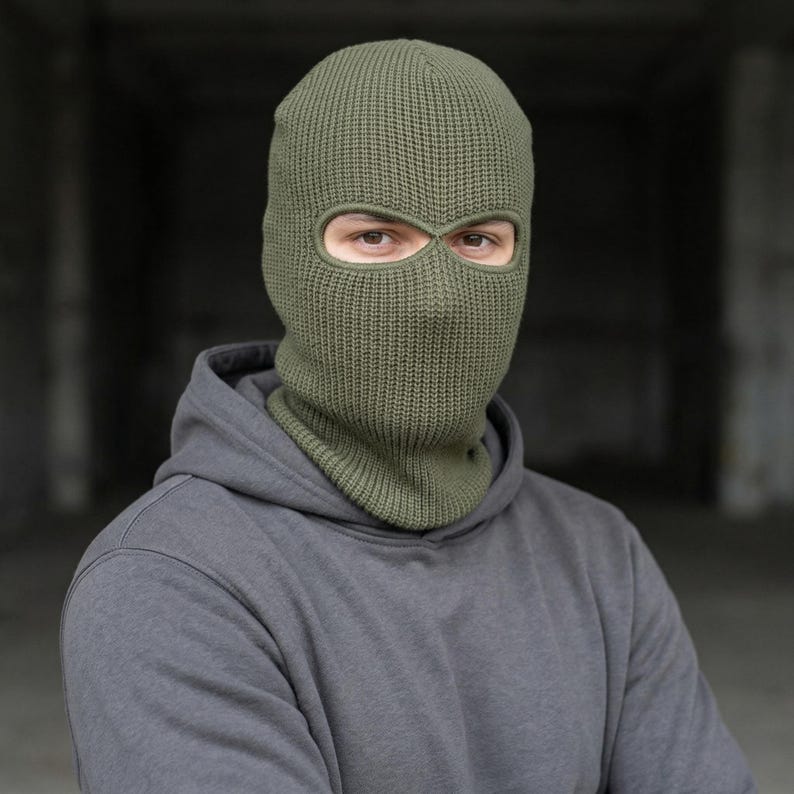 May include: Olive green knitted balaclava with eye and mouth openings. The balaclava is made of a ribbed knit fabric and covers the head, neck, and face, leaving only the eyes visible. The person is wearing a gray hooded sweatshirt.