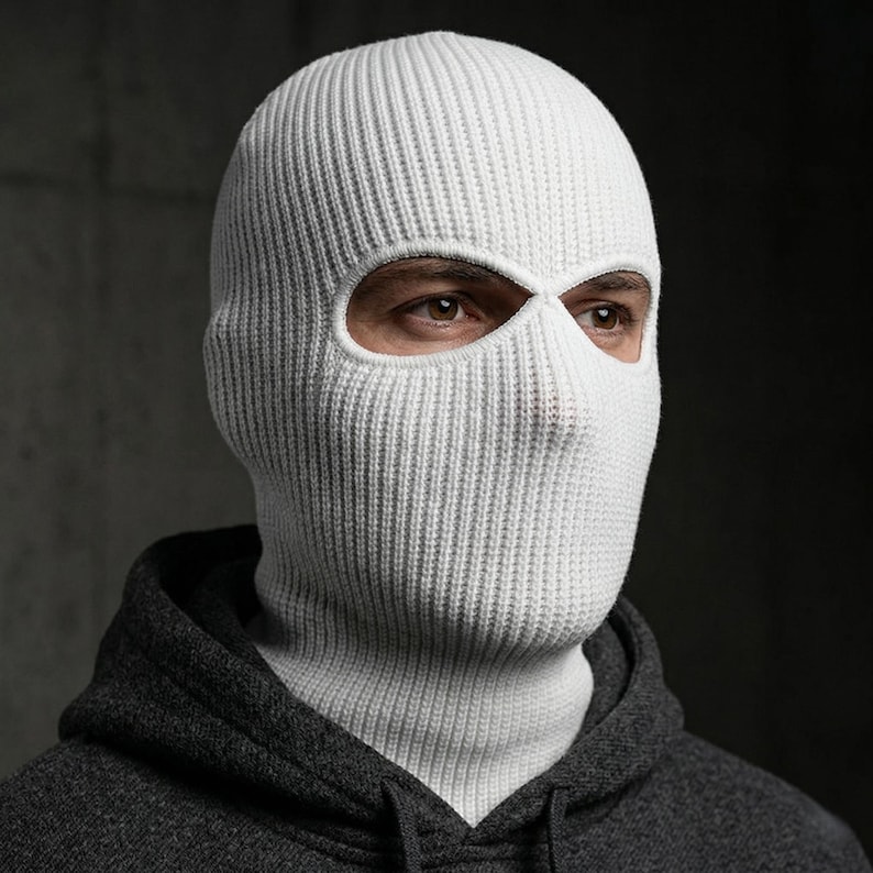 May include: A white knitted balaclava with eye and mouth openings. The head covering is made of a ribbed knit fabric. The person wearing the balaclava is also wearing a gray hooded sweatshirt.