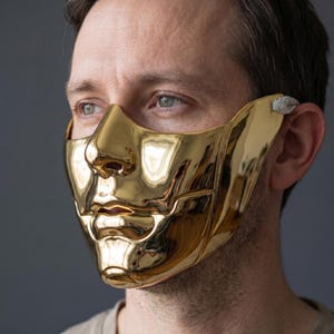 May include: A polished gold mask shaped like a human face. The mask covers the nose, mouth, and cheeks, with a smooth, reflective surface. It has a small, textured attachment near the ear.