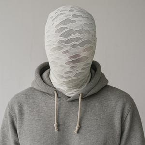 May include: A person wearing a gray hoodie with a white, patterned face covering. The face covering has a distressed, torn design. The hoodie has a drawstring and a kangaroo pocket.