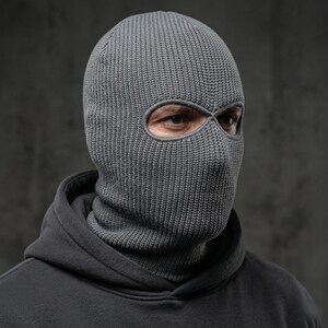 May include: A gray knitted balaclava with eye openings. The head covering is made of a ribbed knit fabric and covers the head and neck. The person is wearing a gray hooded sweatshirt.