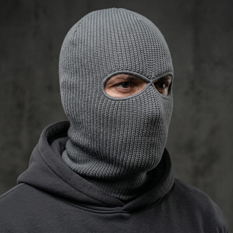 May include: A gray knitted balaclava with eye openings. The head covering is made of a ribbed knit fabric and covers the head and neck. The person is wearing a gray hooded sweatshirt.