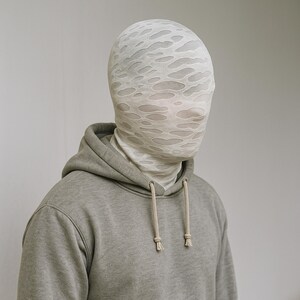 May include: A person wearing a light gray hoodie with a drawstring. The person is also wearing a white head covering with a pattern of irregular holes.