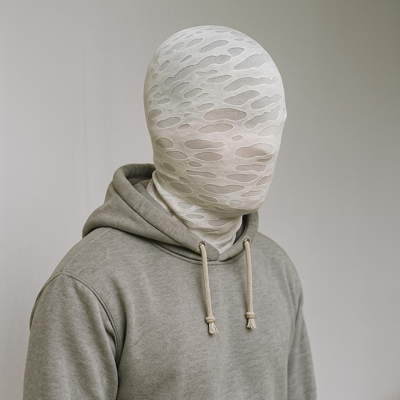 May include: A person wearing a light gray hoodie with a drawstring. The person is also wearing a white head covering with a pattern of irregular holes.