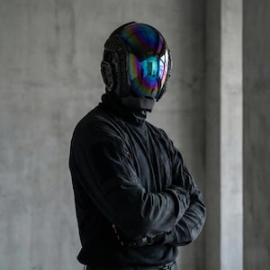 May include: A futuristic black helmet with a reflective, rainbow-colored visor. The helmet has intricate details and covers the entire head. The person is wearing a black turtleneck and long-sleeved shirt, with arms crossed.