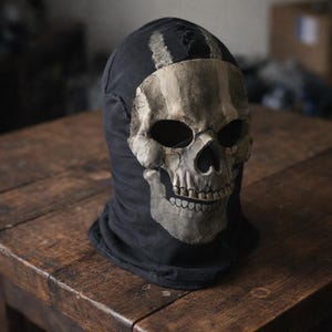 May include: A black balaclava with a distressed skull design on the front. The skull is off-white with gray shading, and the balaclava is resting on a wooden surface. The skull design covers the face area.