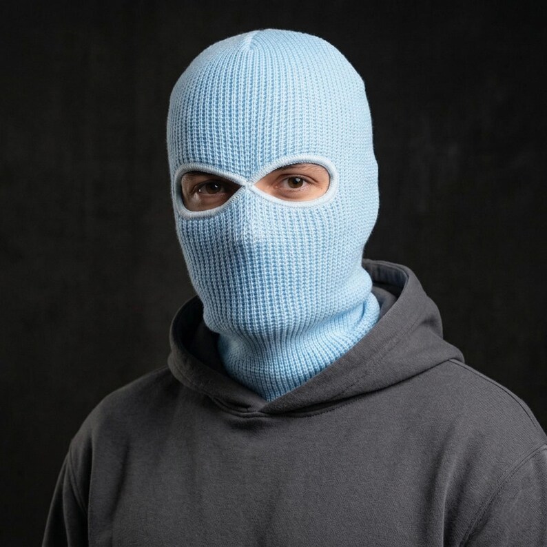 May include: A light blue knitted balaclava with eye and mouth openings. The balaclava covers the head and neck, and is worn with a gray hooded sweatshirt. The background is a dark gray.
