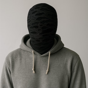 Puede incluir: Una persona con una sudadera gris y una máscara negra. La máscara tiene un diseño texturizado con formas recortadas irregulares. La sudadera tiene un cordón y un bolsillo delantero.