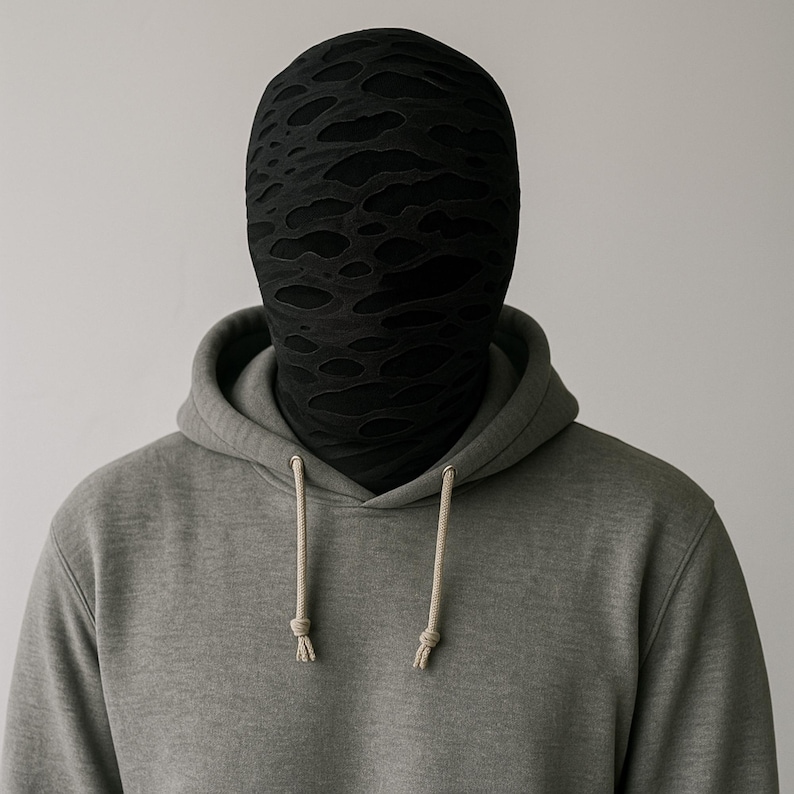 May include: A person wearing a gray hoodie with a black, textured face covering. The face covering has an irregular pattern of cut-out shapes. The hoodie has a drawstring and a front pocket.