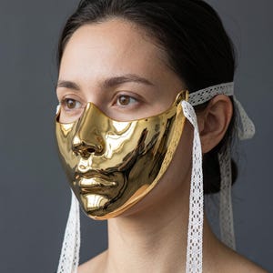 May include: A shiny gold mask shaped like a human face, with a nose and lips. The mask is secured with white lace ties. The mask reflects light, creating a bright, reflective surface. The mask is a decorative item.