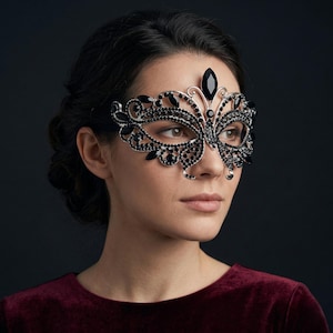 Könnte beinhalten: Eine schwarze und silberne Maske für einen Maskenball mit aufwendigem Filigran und schwarzen Edelsteinakzenten. Die Maske hat ein Schmetterlingsdesign mit einem großen, tropfenförmigen Edelstein auf der Stirn. Die Person trägt ein bordeauxfarbenes Oberteil.