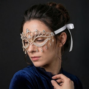 Könnte beinhalten: Eine goldfarbene Maske für einen Maskenball mit klaren Strasssteinen und herabhängenden tropfenförmigen Kristallen. Die Maske wird mit einem weißen Band befestigt. Die Person trägt ein blaues Samt-Oberteil. Die Maske hat ein dekoratives Kronen-Design.