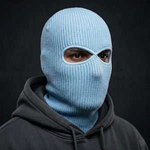 May include: A light blue knitted balaclava with eye and mouth openings. The balaclava is worn over the head and neck, covering the entire face except for the eyes. The person is wearing a gray hooded sweatshirt.