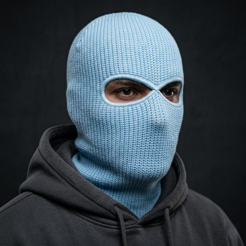 May include: A light blue knitted balaclava with eye and mouth openings. The balaclava is worn over the head and neck, covering the entire face except for the eyes. The person is wearing a gray hooded sweatshirt.