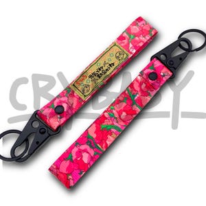 May include: Two keychains with black metal fixings and floral patterns in pink and red. One keychain features a rectangular patch with Japanese text and a cartoon illustration. These keychains are designed for holding keys or small items.