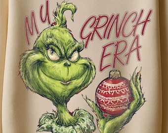 May include: Cream-colored sweatshirt featuring a green Grinch graphic with the text "My Grinch Era" in pink. The Grinch is holding a red and white Christmas ornament. The sweatshirt is hanging on a red hanger.