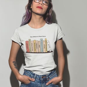 Op de afbeelding: Een wit T-shirt met korte mouwen voorzien van een afbeelding van een boekenplank met diverse boekruggen en de tekst The Villain in Their Story. De boeken zijn gelabeld met titels zoals The Picture of Dorian Gray, American Psycho en Gone Girl.