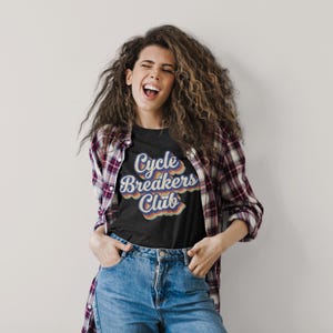 May include: A black t-shirt featuring the text Cycle Breakers Club in a stacked, retro-inspired font with layered outlines in shades of blue, orange, and cream. The shirt is worn under an unbuttoned, long-sleeved, maroon and white plaid flannel shirt.