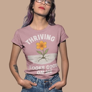 以下が含まれることがあります： 「THRIVING LOOKS GOOD ON ME」の白い文字が入った、くすんだピンク色のTシャツ。黄色い花のグラフィックが特徴です。モデルはブルージーンズとメガネを着用しています。