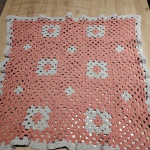 Peut inclure: Une couverture au crochet faite à la main, selon un motif de carrés Granny. La couverture présente un centre rose corail avec des motifs floraux gris et blancs. La bordure est grise et festonnée. La couverture est présentée sur une surface en bois.