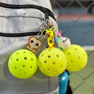 May include: A light gray backpack with a black zipper, holding several keychains. The keychains include two yellow pickleballs, a giraffe, a slice of toast with a smiling face, and a white bunny. The pickleballs have multiple holes.
