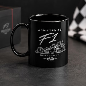 Könnte beinhalten: Eine schwarze Keramiktasse mit einer weißen Grafik eines Formel-1-Rennwagens. Der Text lautet ADDICTED TO F1, SPEED IS A LIFESTYLE, und enthält zwei kleine Zielflaggen am unteren Rand. Das Design ist in einem klaren, weißen Stil aufgedruckt.