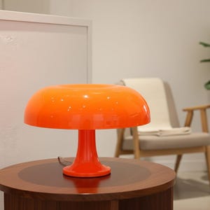 Peut inclure: Lampe de table orange en forme de champignon, avec un abat-jour arrondi et un pied en forme de piédestal. La lampe est posée sur une table en bois brun foncé. En arrière-plan, un mur blanc, une chaise et une plante en pot.