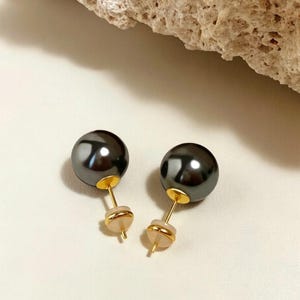 Può includere: Un paio di orecchini a lobo con perle grigio scuro. Le perle sono rotonde e hanno una finitura lucida. I perni e i supporti sono in metallo color oro. Gli orecchini sono esposti su una superficie bianca.