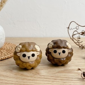 May include: Two small, round, carved wooden sheep figurines with smooth, dark brown surfaces, cream-colored horns, and simple black painted eyes and noses. The sheep are positioned on a light wood surface next to a white ceramic vase with dried flowers.