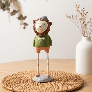 May include: A whimsical wooden lion figurine with a brown mane, wearing a green sweater, orange shorts, and a gray hat. The lion stands on long, striped legs and a gray base. The figurine is placed on a woven mat.