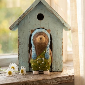 Könnte beinhalten: Eine Holz-Bärenfigur mit blauer Haube und grünen Latzhosen, die in einem hellblauen Vogelhaus steht. Der Bär hat braunes Fell und ist detailliert geschnitzt. Das Vogelhaus hat ein antikes Finish.