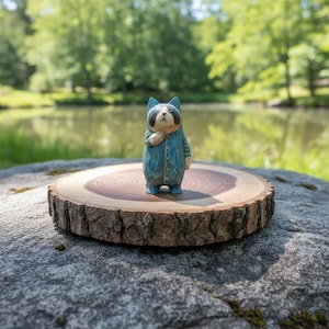 May include: A hand-carved wooden raccoon figurine wearing a blue jumpsuit. The raccoon has a black and white face and is standing on a wooden slice. The background features a lake and trees.