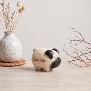 May include: A small, hand-carved wooden figurine of a pug dog with a cream-colored body and black ears and muzzle. The name Cooper is engraved in dark brown letters on the side of the dog. The surface of the wood has a textured, faceted finish.