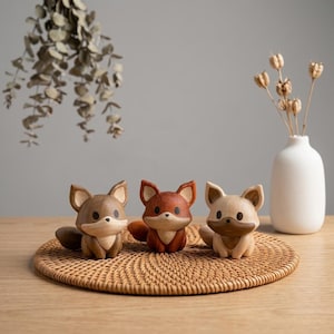 K&ouml;nnte beinhalten: Drei kleine geschnitzte Holzfiguren in Form von F&uuml;chsen, die in einer Reihe angeordnet sind. Der linke Fuchs ist mittelbraun, der mittlere rotbraun und der rechte hellbeige mit einem dunkelbraunen Gesichtsfleck. Jeder Fuchs hat schwarze Augen.