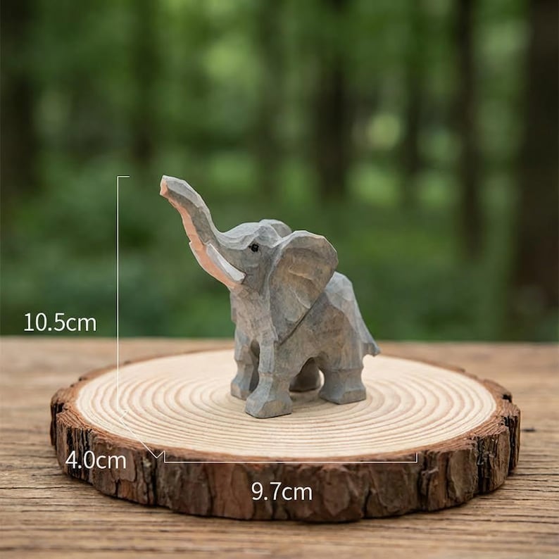 以下が含まれることがあります： 象の木彫りの置物。高さ10.5cm、象牙の牙があり、直径9.7cmの木の輪切りに乗っています。象の鼻は上がっています。木の輪切りの厚さは4.0cmです。背景はぼやけた緑色です。
