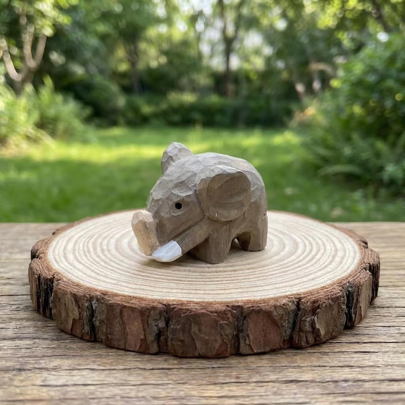 K&ouml;nnte beinhalten: Eine kleine, handgeschnitzte Holz-Elefantenfigur mit hellbraunem Finish. Der Elefant hat detaillierte geschnitzte Merkmale und einen wei&szlig;en Sto&szlig;zahn. Er sitzt auf einer runden, nat&uuml;rlichen Holzscheibe mit sichtbaren Jahresringen.