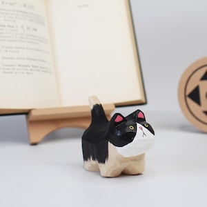 May include: A wooden cat figurine, hand-carved and painted in black, white, and tan. The cat has pink accents on its ears and yellow eyes. The figurine is standing upright, with its tail raised. A unique home decor item.