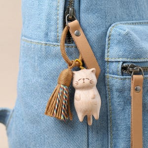 Op de afbeelding: Een lichtgekleurde houten kattenhanger met een kwast en leren riem, bevestigd aan een blauwe denim rugzak. De kattenhanger heeft ge&euml;tste gezichtskenmerken en een kleine staart. De kwast is bruin met kleurrijke draden.
