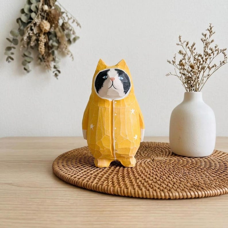 May include: A carved wooden cat figurine wearing a yellow jumpsuit with white star accents. The cat has a black and white face and is standing on a woven mat. A small white vase with dried flowers is in the background.