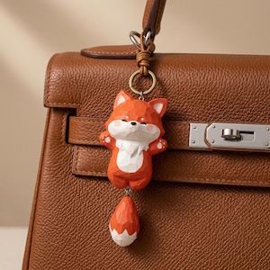May include: A carved wooden fox keyring with a faceted orange and white finish, featuring a separate dangling tail piece. The fox has a small, rounded body, pointed ears, and a white chest patch, attached to a metal ring on a brown leather handbag.