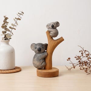 May include: A wooden koala bear sculpture featuring two koalas on a tree branch. The koalas are grey with black eyes, and the tree and base are made of light brown wood. The sculpture is a decorative item.