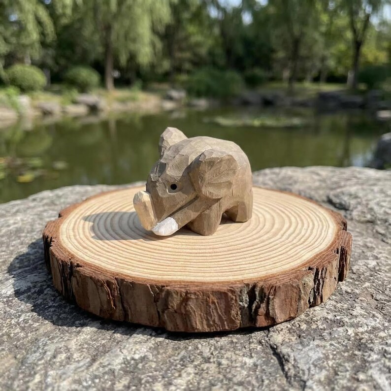 K&ouml;nnte beinhalten: Eine kleine, geschnitzte Holz-Elefantenfigur mit einem wei&szlig;en Sto&szlig;zahn, die auf einer nat&uuml;rlichen Holzscheibe ruht. Der Elefant ist hellbraun mit geschnitzten Details. Die Holzscheibe hat einen rauen Rand.