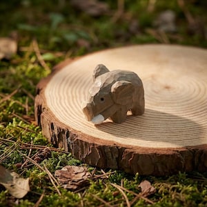 K&ouml;nnte beinhalten: Eine kleine, handgeschnitzte Holz-Elefantenfigur mit hellbraunem Finish. Der Elefant hat detaillierte Merkmale, einschlie&szlig;lich Ohren, R&uuml;ssel und Sto&szlig;z&auml;hne. Er steht auf einer Holzscheibe, umgeben von Moos und Bl&auml;ttern.