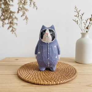 May include: A hand-carved wooden cat figurine wearing a blue onesie with white star accents. The cat has a black and white face and is standing on a round, woven coaster. The figurine is a decorative item.