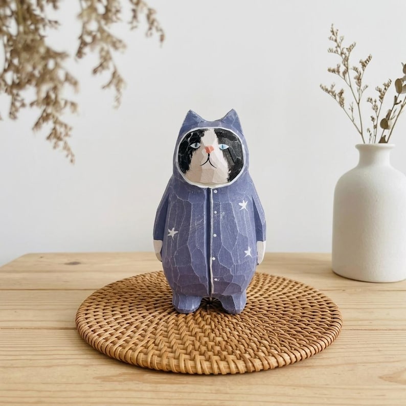 May include: A hand-carved wooden cat figurine wearing a blue onesie with white star accents. The cat has a black and white face and is standing on a round, woven coaster. The figurine is a decorative item.