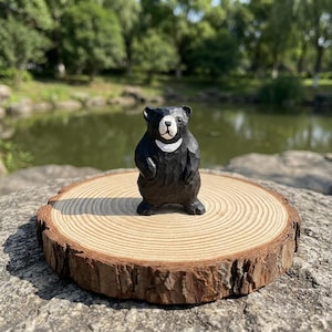 Peut inclure: Figurine d'ours en bois sculpté à la main, corps noir et marque blanche sur la poitrine. L'ours est posé sur une tranche de bois ronde et naturelle, avec des cernes visibles. En arrière-plan, un étang et des arbres flous.