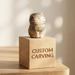 Può includere: Una statuetta di gatto in legno con segni a strisce è appoggiata su un cubo di legno. Il cubo è inciso con le parole "CUSTOM CARVING". Il gatto è scolpito con dettagli e l'estetica generale è naturale e artigianale.