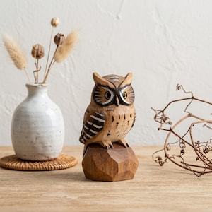 May include: A hand-carved wooden owl figurine with brown, black, and white painted details, perched on a faceted wooden base. The owl features large yellow eyes, dark brown facial markings, and striped wings, showcasing visible wood grain textures.