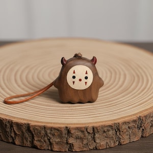 May include: A brown wooden figurine of a cartoon character with red horns and a leather strap. The face features a white circle with black dots and red accents. The figurine rests on a wooden slice, showcasing a natural wood grain.